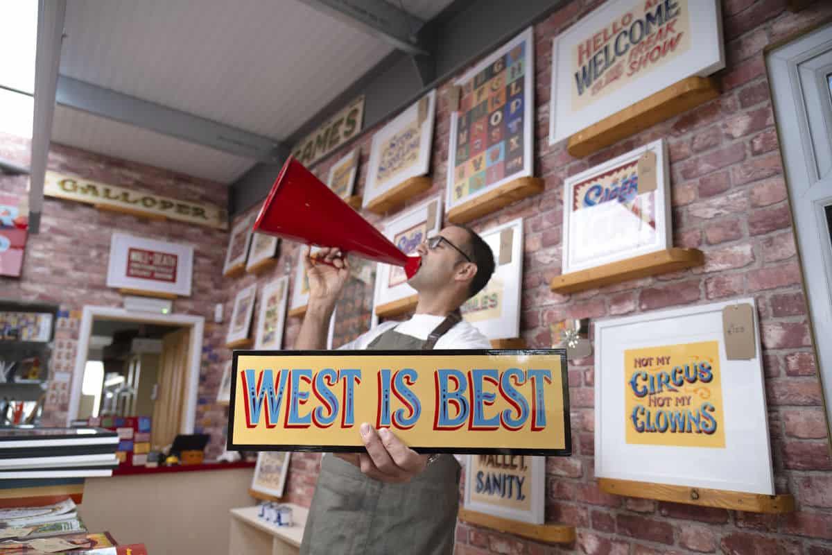 Joby Carter holding a handpainted sign saying West is best in his workshop in Somerset